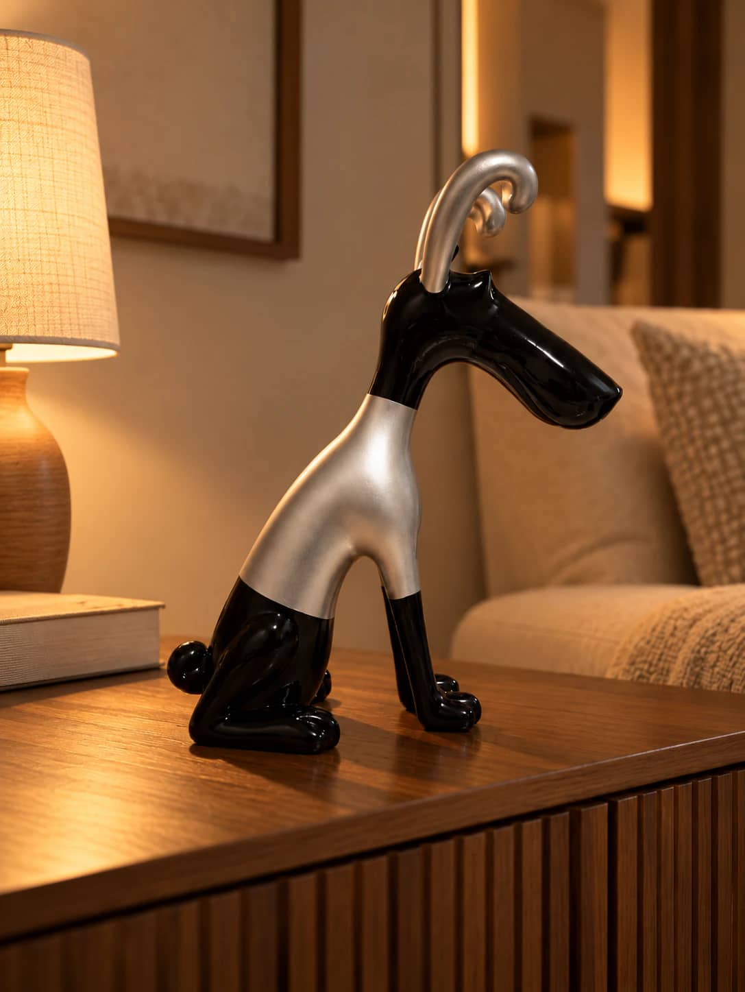 Statuette de chien stylisé noir et argent assise sur un buffet en bois, près d’une lampe allumée